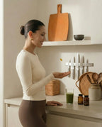 Minimalistische Lifestyle-Szene einer Frau in neutraler Activewear, die in einer warmen Küche ein Supplement zusammen mit einem grünen Drink einnimmt.